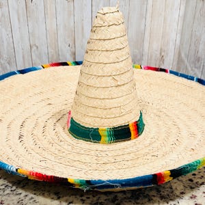 May include: A large, woven straw sombrero with a wide brim and a tall, conical crown. The brim is trimmed with a colorful, patterned fabric in red, blue, yellow, and green. The hat is a traditional Mexican style.