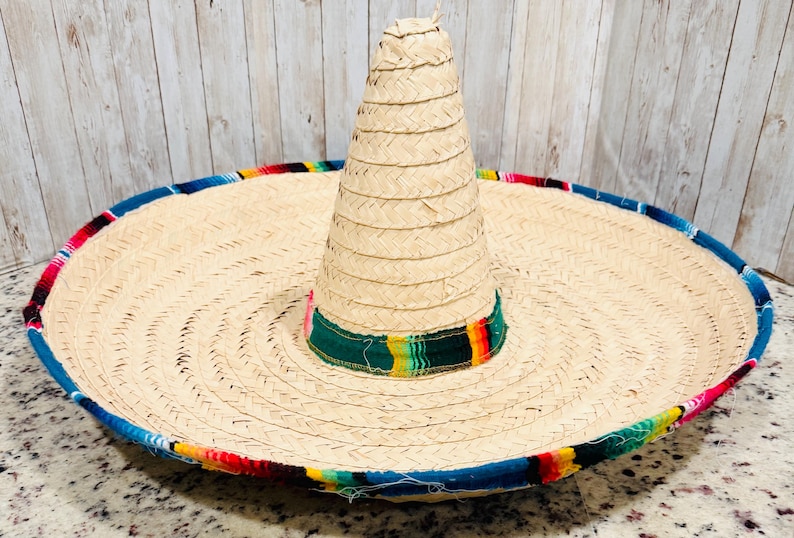 May include: A large, woven straw sombrero with a wide brim and a tall, conical crown. The brim is trimmed with a colorful, patterned fabric in red, blue, yellow, and green. The hat is a traditional Mexican style.
