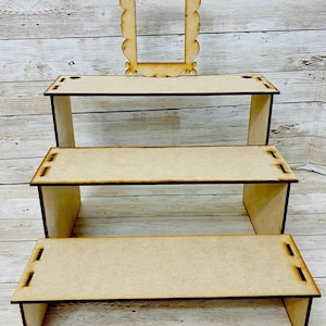 May include: A three-tiered, light brown wooden display stand with a decorative frame on top. The stand has a stepped design, with each level slightly smaller than the one below. The frame is ornate and complements the stand's rustic aesthetic. Ideal for showcasing small items.