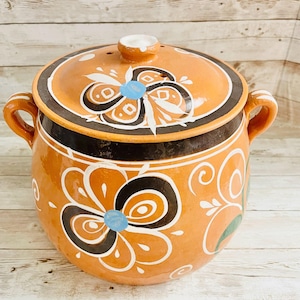 May include: A ceramic cooking pot with a lid, featuring a vibrant orange base and lid. The pot is adorned with floral patterns in black, white, and blue. It has two handles and a small knob on the lid. The pot is likely handmade.