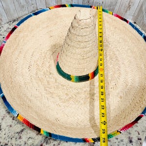 May include: A large, woven straw sombrero with a colorful striped band and trim. The hat measures approximately 26 inches in diameter. The brim is trimmed with blue, red, yellow, and green fabric.