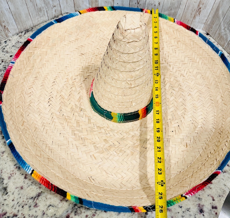 May include: A large, woven straw sombrero with a colorful striped band and trim. The hat measures approximately 26 inches in diameter. The brim is trimmed with blue, red, yellow, and green fabric.