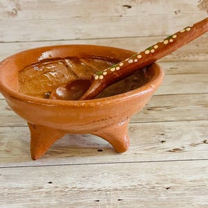 May include: A terracotta molcajete, a traditional Mexican mortar and pestle, filled with a dark sauce. A matching wooden spoon with white and green dots rests inside the bowl. The molcajete has three sturdy legs.