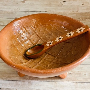 May include: A handmade terracotta bowl with a diamond pattern and three small feet. A wooden spoon with white flower designs rests inside the bowl. The spoon has a green painted interior. This rustic bowl and spoon set is perfect for serving.