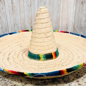 May include: A large, woven straw sombrero with a wide brim and a tall, conical crown. The brim is trimmed with a colorful band of blue, red, yellow, and green stripes. The hat is a traditional Mexican design.