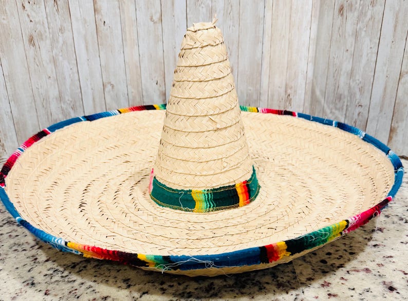May include: A large, woven straw sombrero with a wide brim and a tall, conical crown. The brim is trimmed with a colorful band of blue, red, yellow, and green stripes. The hat is a traditional Mexican design.