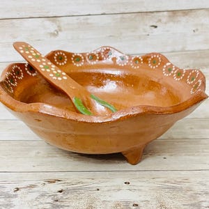 May include: A handmade terracotta bowl with a matching spoon. The bowl has a scalloped rim with white and green painted floral accents. The spoon rests inside the bowl. The bowl is supported by three small feet.
