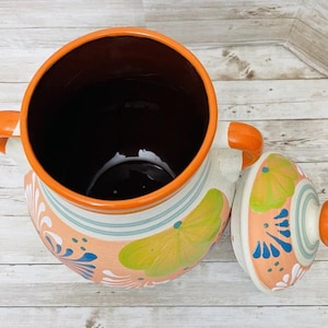 May include: A ceramic pot with a lid, featuring a vibrant design. The pot has a dark interior, orange handles, and a lid with a matching orange knob. The exterior is decorated with a peach, blue, green, and white pattern. The pot is likely used for cooking or serving.