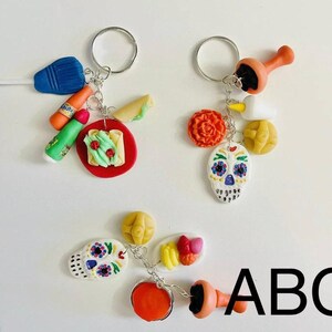 May include: Three keychains with silver rings and various charms. One keychain has a blue popsicle, orange bottle, green tube, and a red plate with food. Another has a skull, orange flower, and yellow ball. The third has a skull, orange flower, and a plate of food. The text "ABC" is visible.