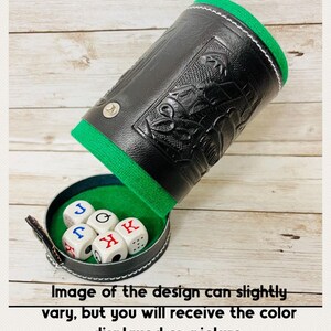 May include: A black leather dice cup with a green felt interior and a decorative embossed design. The cup is open, revealing six dice with letters and symbols. The text at the bottom says "Image of the design can slightly vary, but you will receive the color displayed on picture."