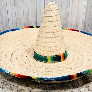 May include: A large, woven straw sombrero with a wide brim and a tall, conical crown. The brim is trimmed with a colorful band of blue, red, yellow, and green. The hat is a traditional Mexican design.