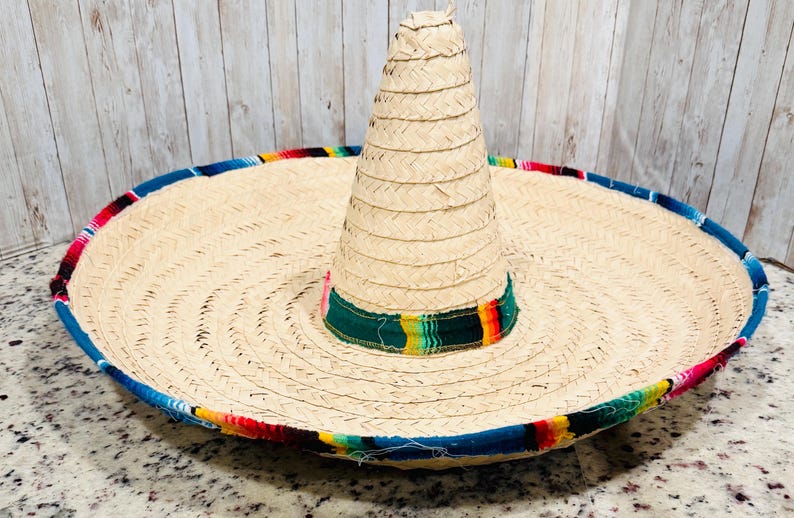 May include: A large, woven straw sombrero with a wide brim and a tall, conical crown. The brim is trimmed with a colorful band of blue, red, yellow, and green. The hat is a traditional Mexican design.