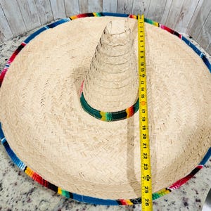 May include: A large, woven straw sombrero with a colorful striped trim. The hat measures approximately 25 inches in diameter. The brim is trimmed with blue, red, yellow, and green fabric.