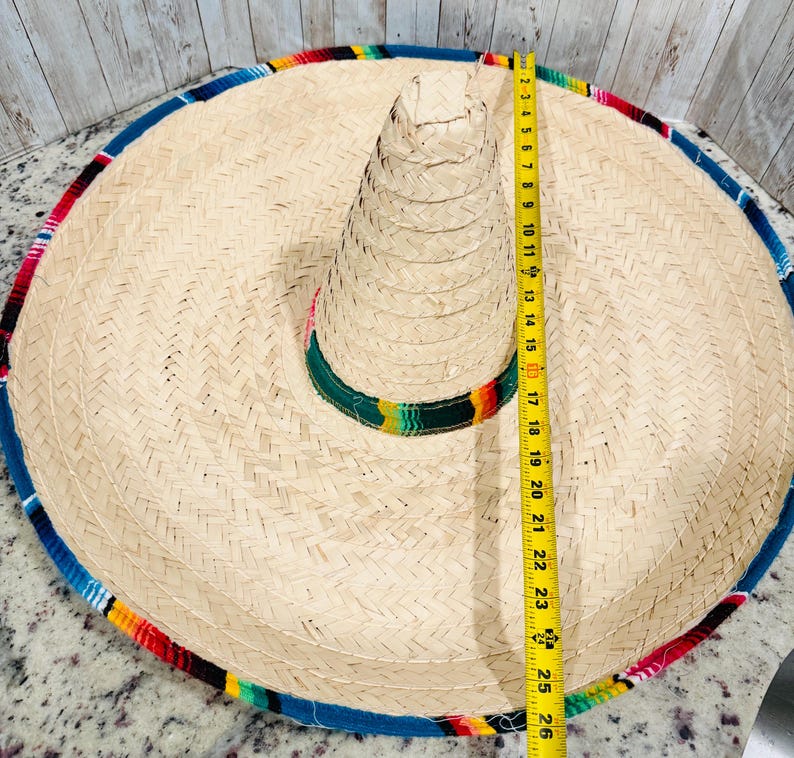 May include: A large, woven straw sombrero with a colorful striped trim. The hat measures approximately 25 inches in diameter. The brim is trimmed with blue, red, yellow, and green fabric.