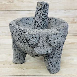 May include: A gray molcajete and tejolote set, made of volcanic rock. The mortar has a bowl shape with three legs and a textured surface. The pestle is cylindrical and fits inside the bowl.