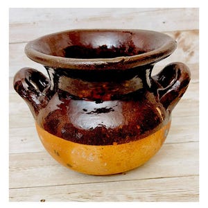 May include: A handcrafted, rustic ceramic pot with a wide rim and two handles. The pot features a rich, dark brown glaze on the upper portion, transitioning to a lighter, tan-colored base. This earthenware vessel is perfect for kitchen decor.