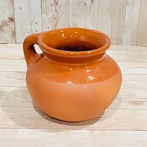May include: A handmade, terracotta-colored ceramic pitcher with a rounded body and a small handle. The pitcher has a glossy finish on the upper portion and a matte finish on the lower portion. The interior is dark, and the pitcher sits on a light-colored wooden surface.