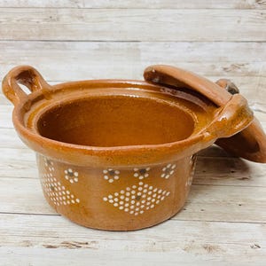 May include: A brown ceramic cooking pot with a lid. The pot has two handles and is decorated with white dotted patterns. The lid rests on the side of the pot. The pot is likely used for cooking and serving food.