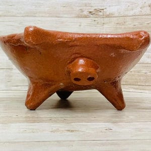 May include: A brown ceramic pig-shaped bowl with three legs. The bowl has a pig snout on the front and two ears on the sides. The bowl is sitting on a wooden surface.