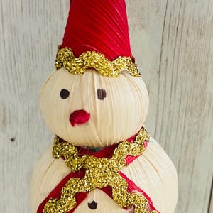 May include: A handmade snowman ornament with a red pointed hat and a gold trim. The snowman has a cream-colored body, black eyes, a red mouth, and a gold scarf. The ornament is suspended by a gold string.