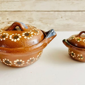 May include: Two terracotta clay pots with lids. The larger pot has blue accents on the handles and a floral pattern with white and brown details. The smaller pot has green accents and a similar floral design. Both pots are on a white surface.