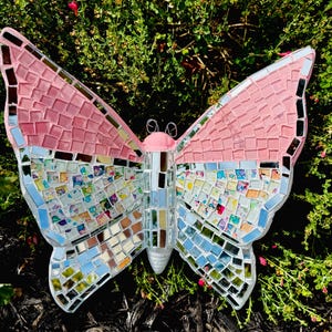 May include: A large, mosaic butterfly sculpture with pink and mirrored wings. The top portion of the wings is pink, while the bottom half is a mix of mirrored and colorful tiles. The butterfly has a pink body and delicate wire antennae.