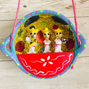 May include: A colorful, handcrafted Day of the Dead decoration featuring three skeleton musicians in yellow hats and white outfits. The musicians are inside a blue and red decorative bowl with a gold glitter background, suspended by a pink string.