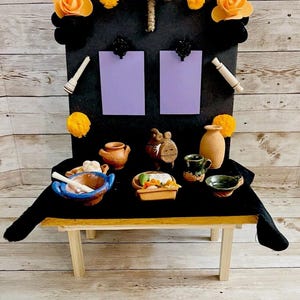 May include: A miniature Day of the Dead altar with a black backdrop, adorned with orange flowers, pom-poms, and a woven cross. The altar features small ceramic vessels, a mortar and pestle, and a miniature table.