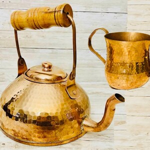 May include: A copper tea kettle and a copper pitcher with a hammered texture. The kettle has a wooden handle and a spout. The pitcher has a curved handle. Both items are displayed on a light wood surface.