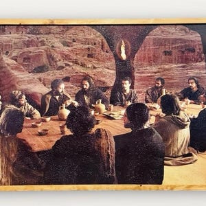 May include: A framed painting depicting a gathering of people around a table. The artwork features a warm color palette with figures in dark clothing. The background shows a landscape with architectural elements, and the frame is a light brown color.