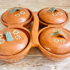 May include: A terracotta serving dish with four bowls and lids. Each lid has a teal handle and white and teal floral accents. The bowls are connected by a handle, and the piece is decorated with white and teal floral patterns.