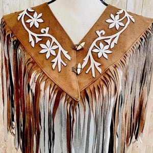 May include: Brown suede fringed vest with white floral cutouts and decorative buttons. The vest has a V-neck collar and long fringe detailing along the bottom and sides. The vest is displayed on a mannequin.