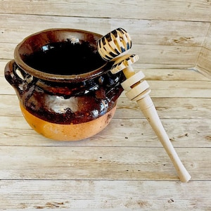 May include: A handmade, brown ceramic pot with a terracotta base and two handles. A wooden molinillo, with a carved top and a long handle, rests inside the pot. The molinillo is used to froth hot chocolate.