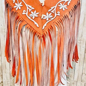 May include: Orange suede fringed shawl with white floral applique. The shawl has a V-neck collar and is fastened with two wooden buttons. The fringe is a gradient of orange and peach.