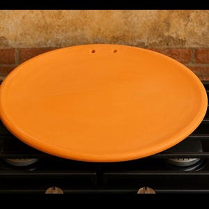 May include: An orange, round, ceramic pizza stone with two small holes near the top edge. The stone is sitting on a black gas stove. The background features a brick wall.