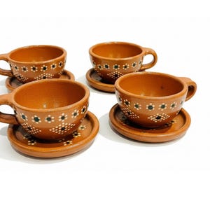 May include: Four terracotta-colored ceramic teacups with matching saucers. Each cup and saucer is adorned with a repeating pattern of white and black floral designs. The cups have rounded handles.