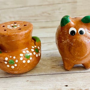 May include: A pair of handmade ceramic salt and pepper shakers. One is a brown, cylindrical shaker with a lid and small holes. The other is a brown pig-shaped shaker with green accents and white eyes. Both are decorated with white dots.