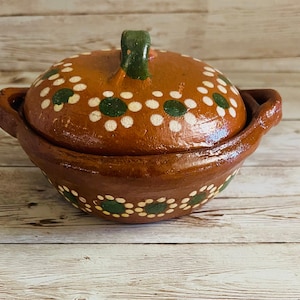 May include: A handcrafted, brown ceramic casserole dish with a lid. The dish features white and green floral patterns and two side handles. The lid has a green handle. This rustic dish is perfect for serving and cooking.
