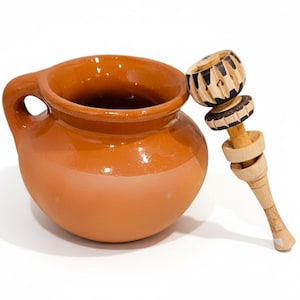 May include: A terracotta clay mug with a handle and a wooden molinillo whisk. The mug is a warm brown color, and the whisk has a carved design. The molinillo is resting against the side of the mug.