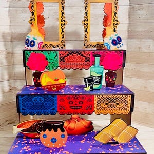 May include: A three-tiered wooden display stand featuring colorful cutouts of skulls, flowers, and food items. The top arch reads BIENVENIDOS, while the bottom tier displays the text DIA DE LOS MUERTOS. The stand is decorated with vibrant papel picado patterns.