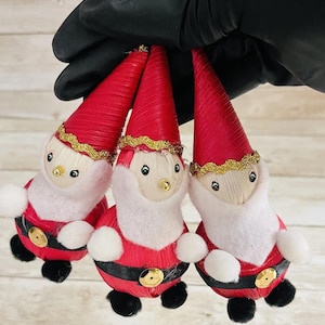 May include: Three Christmas ornaments shaped like Santa Claus figures. Each ornament has a red conical hat, a white beard, and a red suit with a black belt and gold buckle. The ornaments are held against a light-colored background.