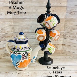 May include: A ceramic pitcher and six matching mugs with a floral design, displayed with a black mug tree. The pitcher and mugs feature blue, orange, and green accents. The text on the image reads "Pitcher 6 Mugs Mug Tree".