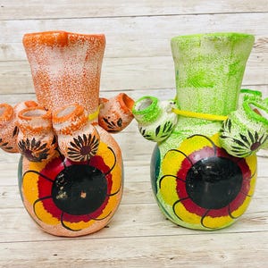 May include: Two hand-painted ceramic vases. One is orange with a white speckled finish, and the other is green. Both have a bulbous base with a floral design in yellow, red, and black. Small, cup-like structures encircle the vase.