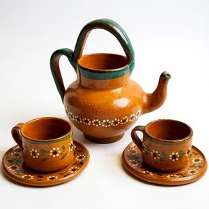 May include: A terracotta-colored tea set with a teapot and two cups and saucers. The teapot has a green handle and rim, and the cups and saucers are decorated with white and green floral patterns. The set is likely handmade.