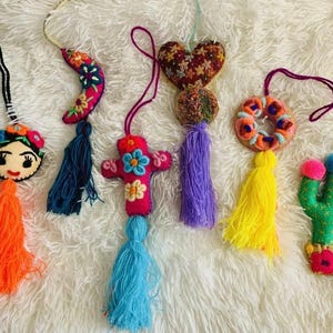 May include: A collection of colorful, handmade fabric ornaments. Designs include a Frida Kahlo face, a crescent moon, a cross, a heart, a wreath, and a cactus. Each ornament has a tassel and a string for hanging.