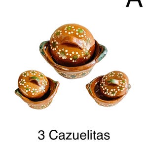 May include: Three brown cazuelitas, or small clay pots, with lids. The pots have a rustic design with white and green accents. The text "3 Cazuelitas" is displayed below the pots. Two pots are 2x3 inches, and one is 3x5 inches.