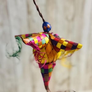 May include: A colorful, woven ornament with a star-like shape. The ornament features a checkered pattern in various colors, including pink, yellow, green, and blue. It is suspended by a brown string and a blue bead.