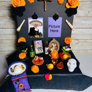 May include: A Day of the Dead altar with a black backdrop, adorned with orange paper flowers, a woven cross, and miniature food items. Includes a photo, the text "Picture Here", and a skull.