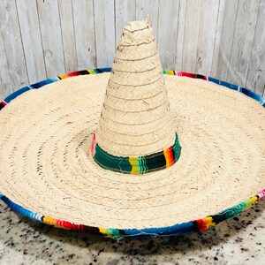 May include: A large, woven straw sombrero with a wide brim and a tall, conical crown. The brim is trimmed with a colorful, patterned fabric in shades of blue, red, yellow, and green. The hat is a traditional Mexican design.