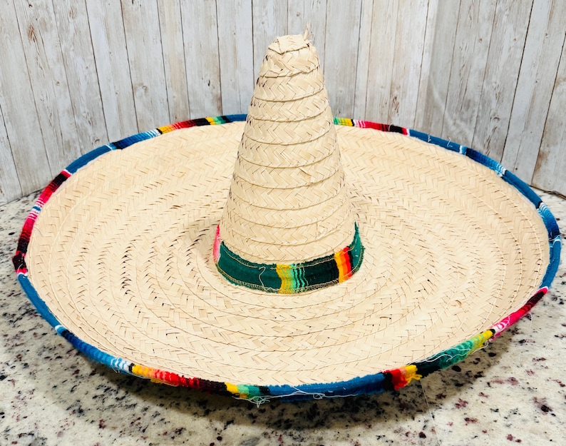 May include: A large, woven straw sombrero with a wide brim and a tall, conical crown. The brim is trimmed with a colorful, patterned fabric in shades of blue, red, yellow, and green. The hat is a traditional Mexican design.